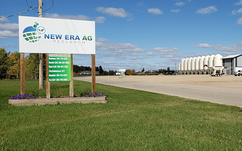 Photograph of the New Era Ag property. Foreground is green grass and a large sign with the New Era Ag logo. Background is the property with road and buildings. Blue sky with clouds.
